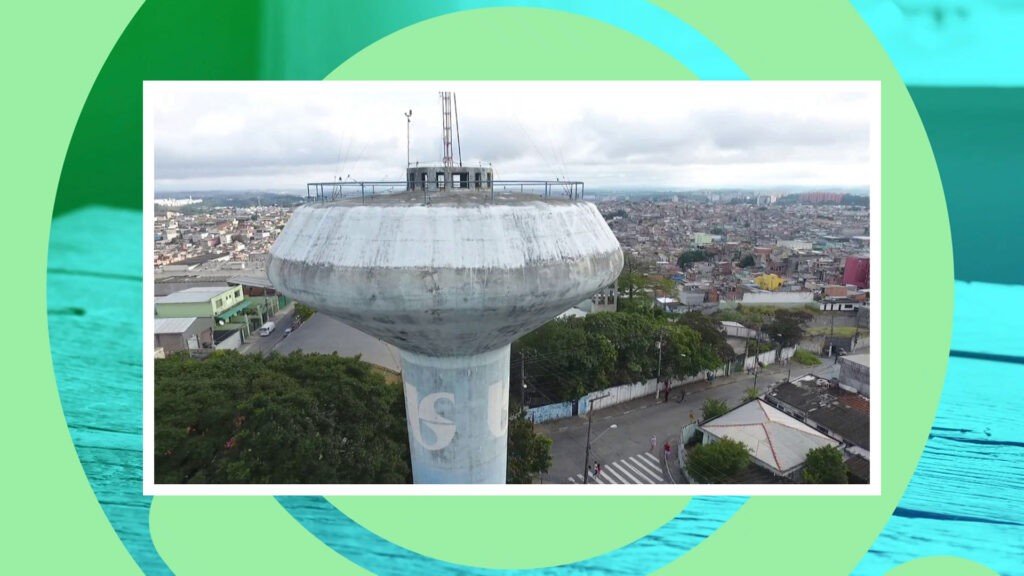 vista aérea de uma caixa d'água da Sabesp na cor azul. Ao fundo se vê o bairro de americanópolis com casas simples. - a foto continua na tela.